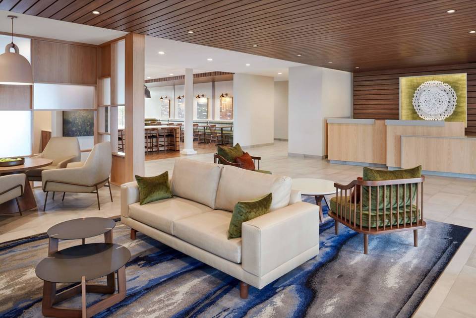 Rendering of the hotel lobby: a cream sofa and wooden armchairs on a blue patterned rug, with the front desk under a wood-slat ceiling